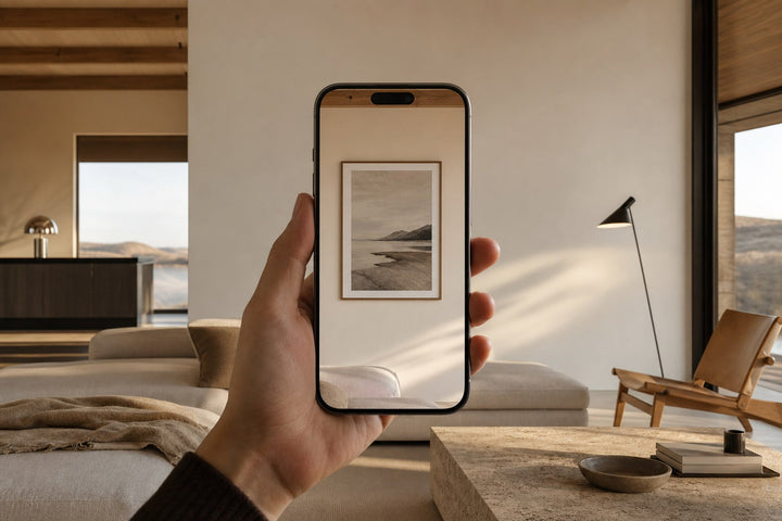 Nordic editorial living room featuring hand holding a modern phone that previews one correctly scaled framed artwork on the empty wall
